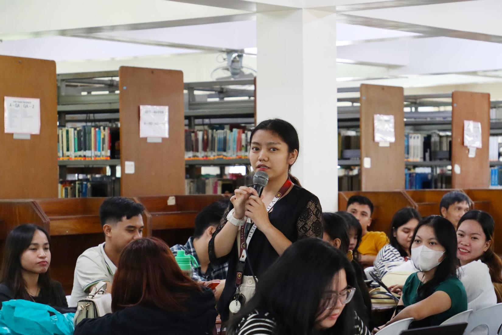A girl student holding a microphone asks a question inside a library during a forum
