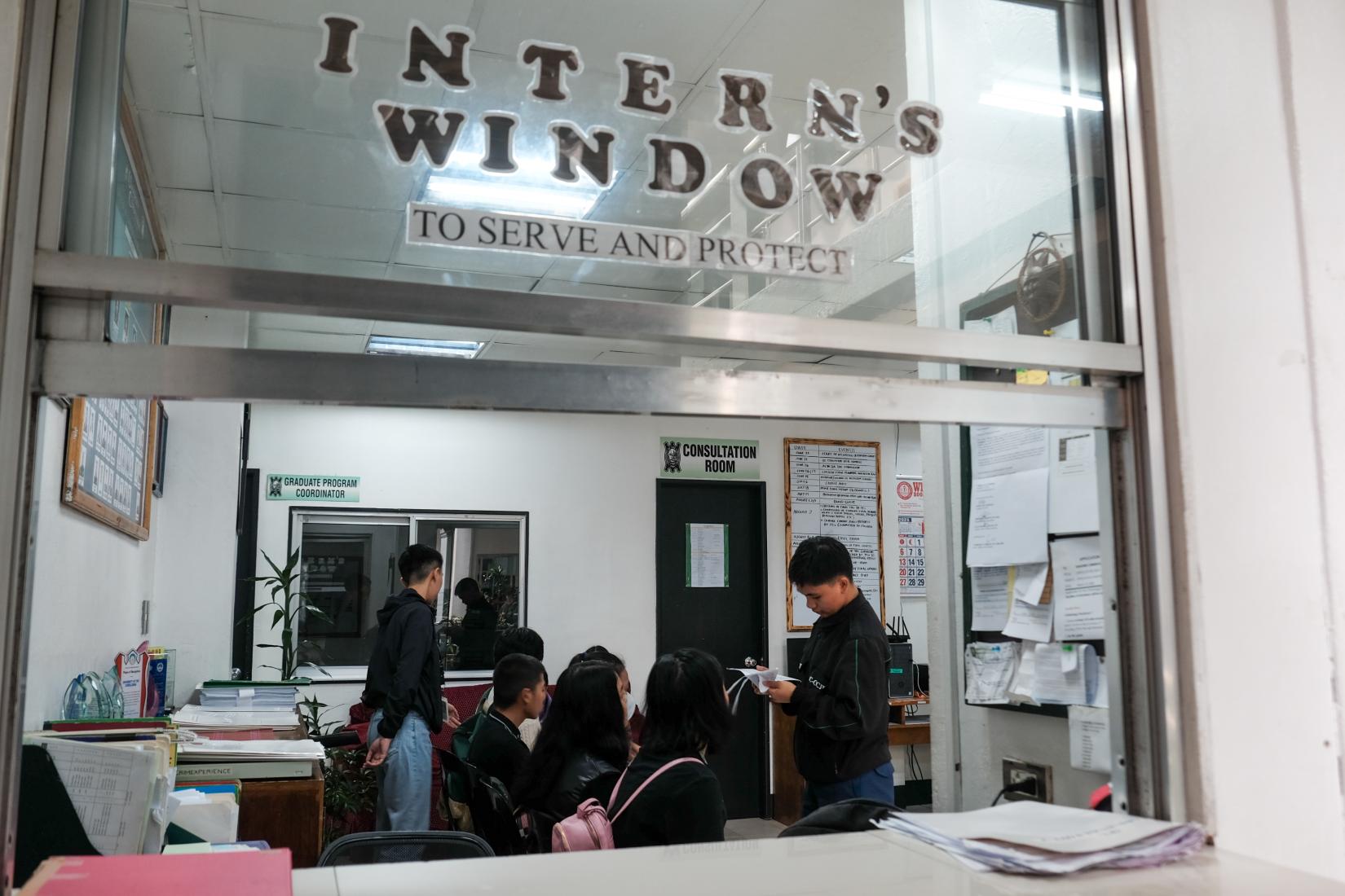 Through a window labelled "Intern's Window", a young student is seen assisting persons seated inside an office